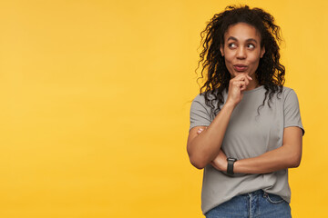 African american girl wears grey t-shit looks aside at copy space with thoughtful facial expression an touching her chin, isolated over yellow background