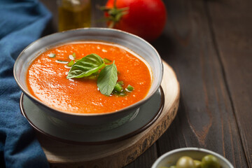 A plate of cold gazpacho made from tomatoes, cucumber peppers with tabasco sauce on a dark wooden table with olive oil and ingredients