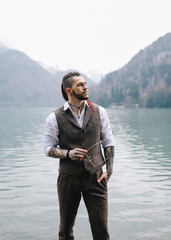 Fototapeta premium A brutal thoughtful young man groom with a dreadlocked hairstyle in a fashionable elegant wedding suit stands on the shore of a lake against the background of misty mountains in nature