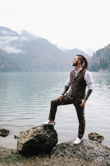 Fototapeta na wymiar A brutal thoughtful young man groom with a dreadlocked hairstyle in a fashionable elegant wedding suit stands on the shore of a lake against the background of misty mountains in nature