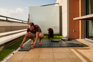 middle-aged man setting up an artificial floor on his terrace with his Labrador retriever sitting next to him