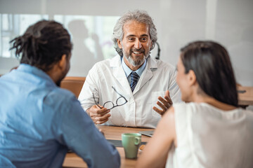 Obraz premium Male doctor offering medical advices to a young couple in office. Husband and wife in a clinic, Beautiful young couple at pregnancy consultation period. Couple meeting medical specialist at hospital