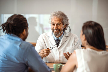Obraz premium Male doctor consulting with young couple in doctor's room. Male doctor explaining diagnosis to a couple during a visit. Smiling doctor looking at happy couple in medical office
