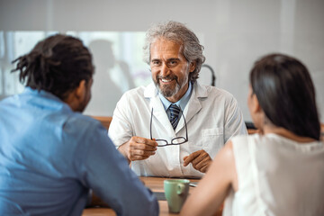 Obraz premium Doctor speaking to young couple. Doctor Using Tablet Computer Discussing Treatment With Patients. Mature Doctor Involved In Serious Discussion With His Patients