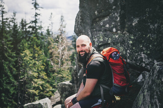 Tourist man on a mountain near large stones watching the natural landscape on a sunny day with his backpack. A handsome lisy bearded man sits and rests on a stone.