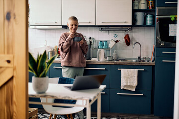 Senior adult woman standing in a kitchen and eating her breakfast.