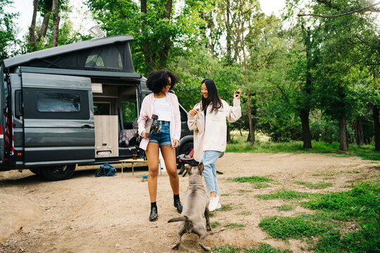 Young diverse women with dog having fun near camper van in forest