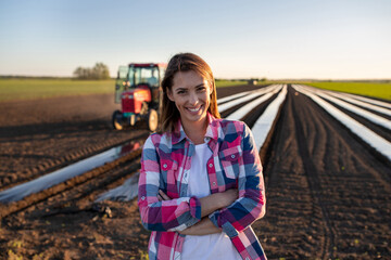 Female agronomist standing in front of field and tractor smiling looking at camera - obrazy, fototapety, plakaty