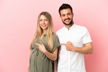 Pregnant woman and man over isolated pink background pointing finger to the side and presenting a product