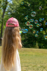 Little girl with pink hat and long hairs blowing soap bubbles in air in front of the forest background. Girl standing back to camera