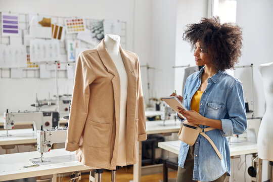 Positive African-American tailor in denim shirt writes in notebook looking at mannequin with jacket in workshop