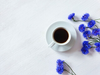 Obraz na płótnie Canvas Coffee cup with saucer and blue cornflowers on white wood table. Copy space, product advertising backdrop or background. Empty place to display product packaging. Showcase mockup. Top view