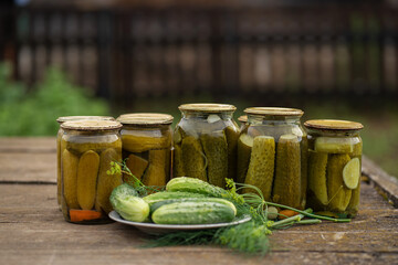 Glass jars with canned cucumbers in a rustic style on a wooden table. Plate with fresh cucumbers and dill