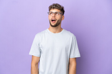 Delivery caucasian man isolated on purple background looking up and with surprised expression