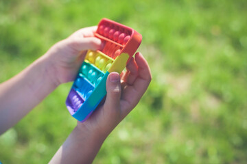 Concept of fidgeting game for stressed child. Pop It sensory antistress. Fidget kid colorful rainbow game on natural background. Child play pop it sensory toy, hands close-up. Stress, anxiety relief