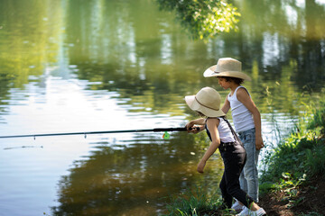 Fototapeta na wymiar A boy and a girl with curly hair in straw hats stand with a fishing rod by the pond. Children fishing on a summer day.
