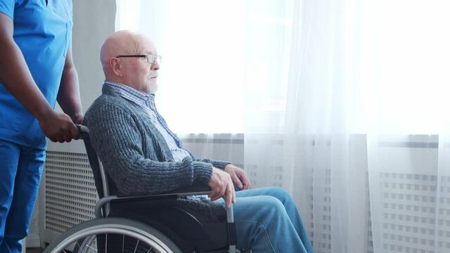 African-American caregiver and old disabled man in a wheelchair. Professional nurse and handicapped patient in a nursing home. Assistance, rehabilitation and health care.