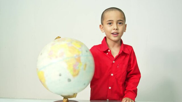 A boy with a skinhead in a red shirt is spinning the globe..A boy with skinhead hair in a red shirt is spinning a globe and .explaining about the world map in a white room..