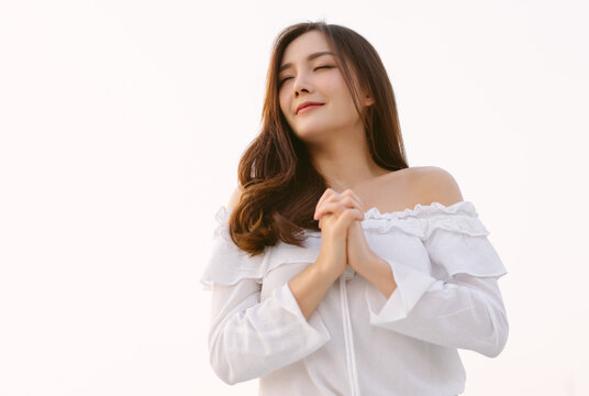 Portrait of beautiful Asian woman with appealing smile and holding clasped hands with closed eyes and grateful gesture on face. Praying or thanking God for everything that she has, with copy space
