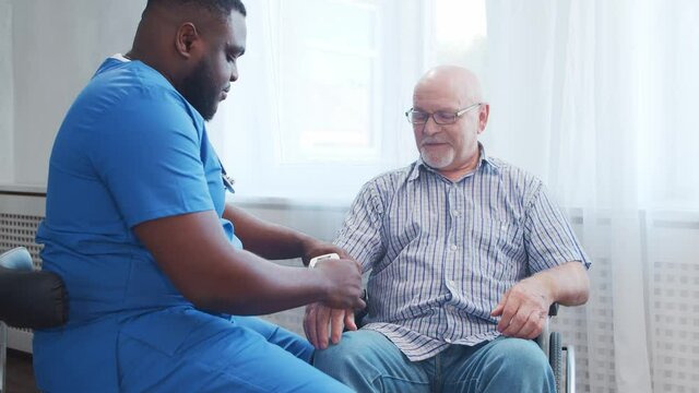 African-American caregiver is measuring disabled old man blood pressure. Professional nurse and handicapped patient in a nursing home. Assistance, rehabilitation and health care.