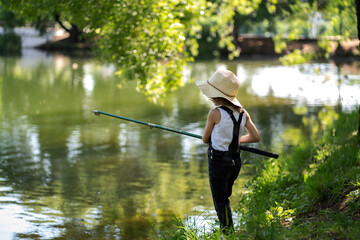 Fototapeta na wymiar A little girl with curly hair in a straw hat and black overalls stands with a fishing rod by the pond. Child fishing on a summer day