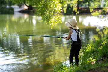 A little girl with curly hair in a straw hat and black overalls stands with a fishing rod by the pond. Child fishing on a summer day © Анжелла Антоновская