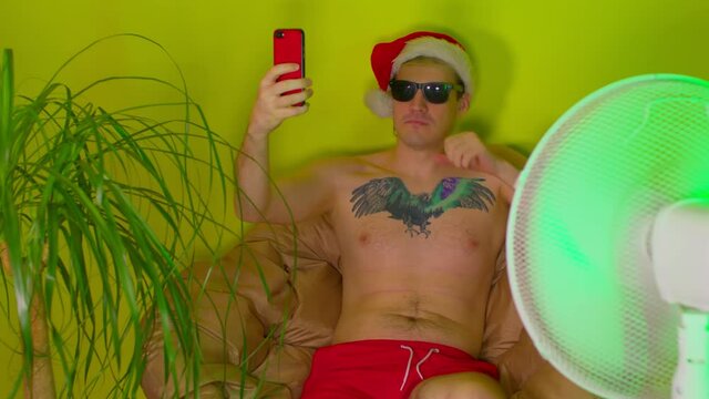 Young man in santa hat with bare torso takes selfie on smartphone, sitting in armchair before fan. Adult guy in sunglasses enjoying his vacation at home as at resort. Concept of isolation.