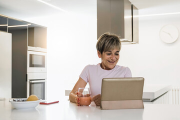 Happy senior woman using digital tablet while eating breakfast indoor at kitchen - Focus on face