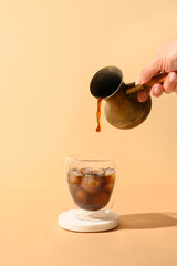 Pouring coffee in glass cup with ice cube on modern beige background. Vertical format.