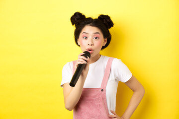 Cute asian teen girl with bright makeup, singing in microphone karaoke, standing against yellow...