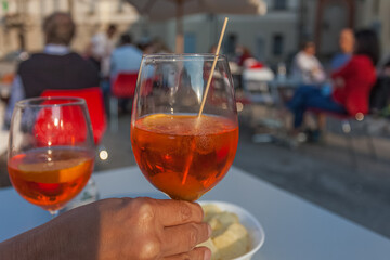Hand holding a glass of spritz with blurred people sitting in a bar in the background, Vittorio Veneto, Italy