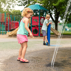 Little happy children playing at water splash pad fountain in park playground on hot summer day.