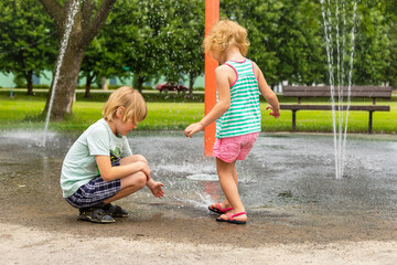 Little happy smiling children playing with water at splash pad in the local public park on hot sunny summer day. Small beautiful kids having fun at fountain playground.
