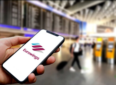 Eurowings Logo wird auf dem Smartphone einer Person angezeigt welche an einem Flughafen vor den Abflugtafeln und Passagieren mit Gepäck steht, Reisen, Airport, Fliegen