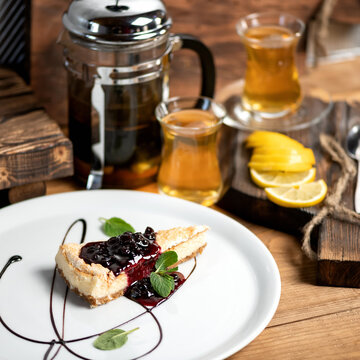 Delicate Dessert. Delicious Cheesecake On White Plate. Piece Of Pie Decorated With Currant Jam And Mint Leaves. Kitchen Table With Glassware Of Tea On It. Soft Focus. Copy Space.