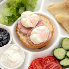 Sandwich with sausage and poached eggs on white portioned plate with vegetables and sauce. Classic European or American Continental breakfast. Close-up shot. Soft focus.