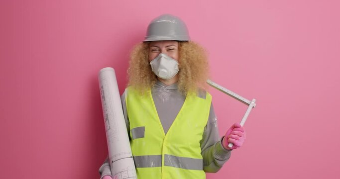Glad curly haired skilled female builder holds blueprint and tape measure came on construction site wears helmet protective respirator and uniform isolated over pink background. Industrial worker