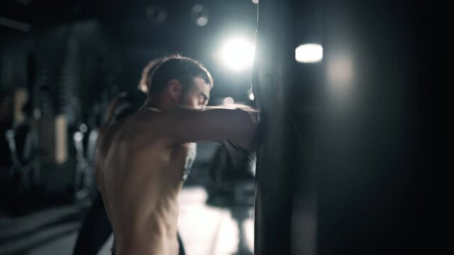 A muscular man and woman are hits punching bag in the gym. Athletes are boxing in dark hall. Self confident people is training indoors. Maximum focus and strength. Concept of health and sports.