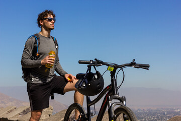 Young biker with sunglasses holding water bottle next to mountain bike on sunny day. Handsome man...