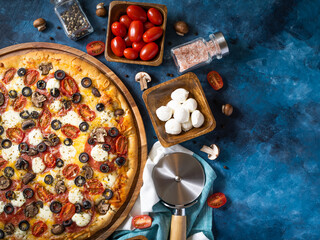 On a blue background on the table lies a pizza beautifully decorated with tomatoes, cheese, olives. Various ingredients are spread around - tomatoes, mushrooms in wooden bowls, a knife.