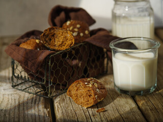 On a rough wooden table, pieces of brown bread in an openwork basket on a brown napkin. There is a glass cup with milk next to it. In the background is a jug of milk. Country style.