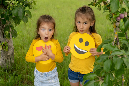 The sisters are holding the faces of happy emoticons in their hands. Two girlfriends in the park under an apple tree look with surprise and perplexity with his mouth open