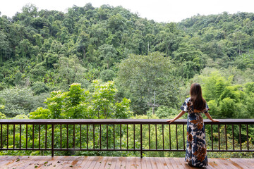 Beautiful woman in long dress standing at wooden balcony look over green mountain view over sky on morning foggy background.