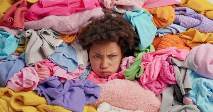 Displeased woman with Afro hair sticks head through pile of multicolored laundry expresses negative emotions cleans out wardrobe collects items for donation. Disorder in closet. Decluttering