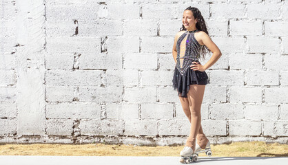Fototapeta na wymiar Full-length portrait of Latina roller skater with curls and competition outfit outdoors smiling on a sunny morning in front of a white block wall