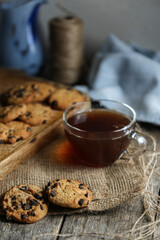 A cup of hot black tea in a glass cup and a teapot press and a gray ceramic plate with cookies with chocolate chips on a wooden tabletop with a board and sackcloth.