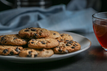 A cup of hot black tea in a glass cup and teapot press and a gray ceramic plate with cookies with chocolate chips on a gray ceramic counter top with a kitchen towel.