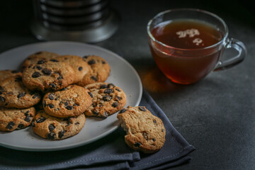 A cup of hot black tea in a glass cup and a teapot press and a gray ceramic plate with cookies with chocolate chips on a gray ceramic counter top with a gray linen napkin.