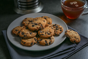 A cup of hot black tea in a glass cup and a teapot press and a gray ceramic plate with cookies with chocolate chips on a gray ceramic counter top with a gray linen napkin.