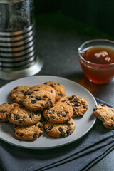 A cup of hot black tea in a glass cup and a teapot press and a gray ceramic plate with cookies with chocolate chips on a gray ceramic counter top with a gray linen napkin.